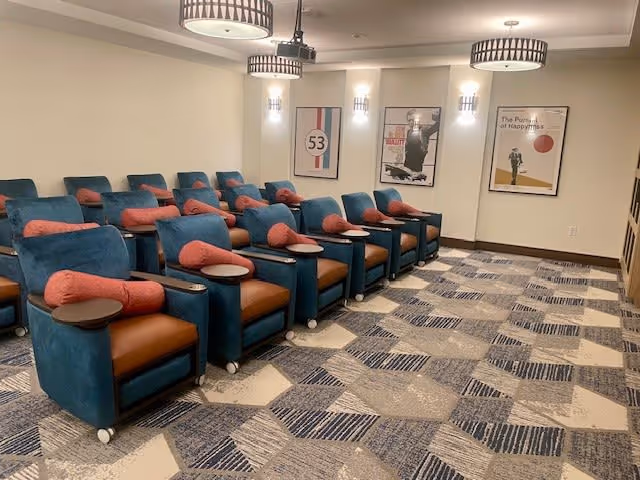 A small theater room with two rows of blue and brown cushioned recliner chairs, each equipped with a small round side table and a coral-colored pillow. The room has patterned carpet flooring, three modern ceiling lights, and three framed posters on the far wall.