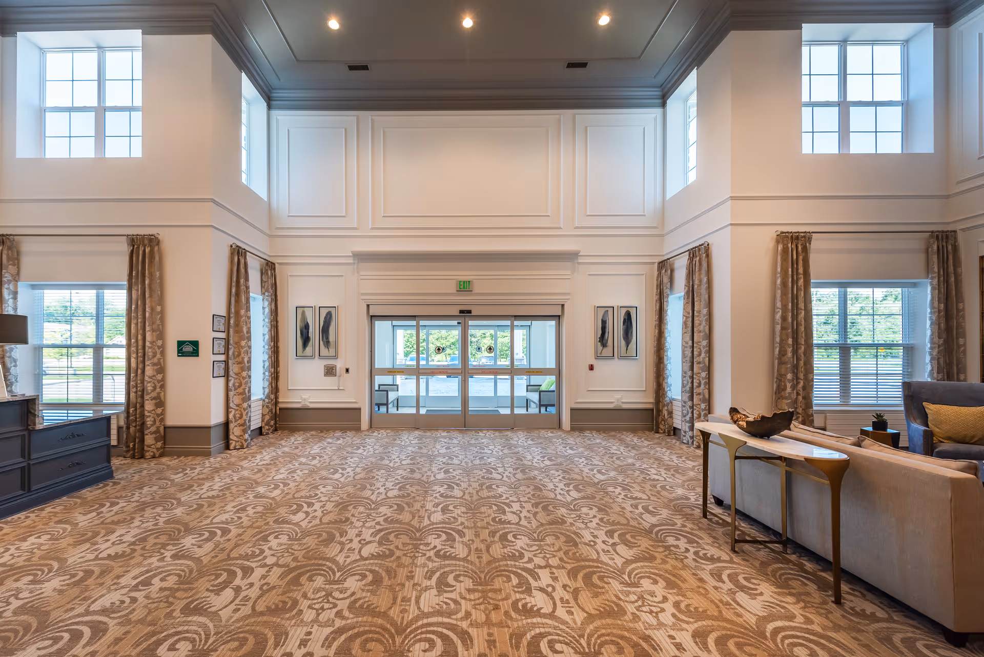 Spacious and elegant lobby area with high ceilings, large windows with curtains, patterned carpet, and a seating area with sofas and a side table. The entrance features glass double doors with an exit sign above.