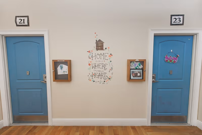 Two blue doors numbered 21 and 23 in a hallway with wooden floors. Between the doors is a wall decoration with the phrase 'Home is where the heart is' surrounded by floral designs and a small house illustration. Each door has a wooden display case mounted on the wall next to it, containing pictures and artwork. The door on the right has a colorful 'Welcome' sign hanging on it.