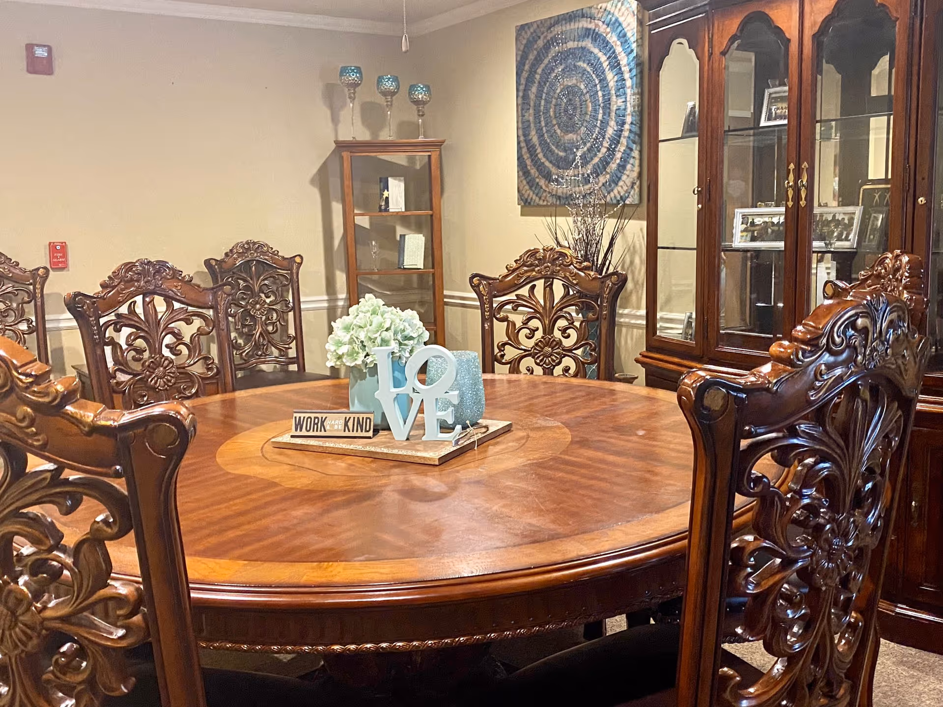 A round wooden dining table with ornate carved wooden chairs surrounding it. On the table is a centerpiece featuring white flowers, a decorative block spelling 'LOVE', and a sign that says 'WORK HARD & BE KIND'. In the background, there is a wooden display cabinet with glass doors and shelves, a small wooden corner cabinet with decorative items on top, and a circular blue and beige wall art piece.