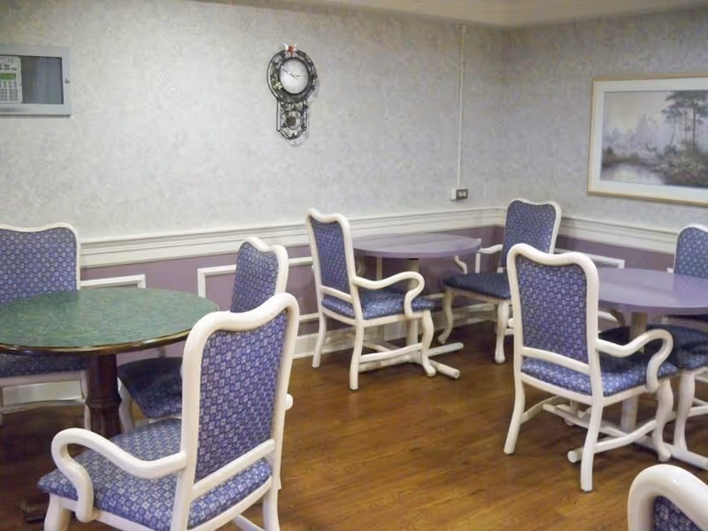 Interior room with several round tables and cushioned chairs with white frames and blue patterned upholstery. The room has wood flooring, light-colored walls with a purple lower section, a wall clock, and a framed landscape painting.