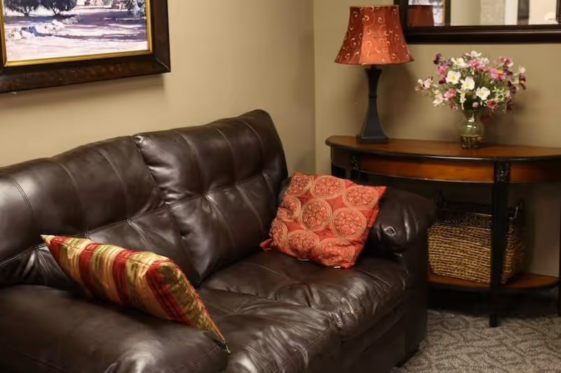 Dark brown leather sofa with patterned throw pillows beside a wooden console table holding a lamp and a vase of flowers.