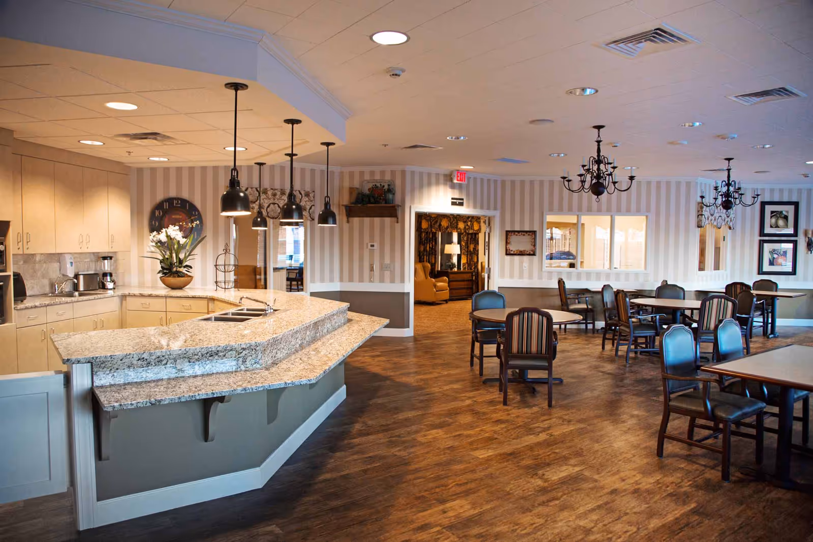 Interior view of a dining area in Creekside Inn Memory Care Community featuring a large granite countertop kitchen island with pendant lights above, multiple round and rectangular dining tables with chairs, wood flooring, striped wallpaper, and chandeliers hanging from the ceiling.
