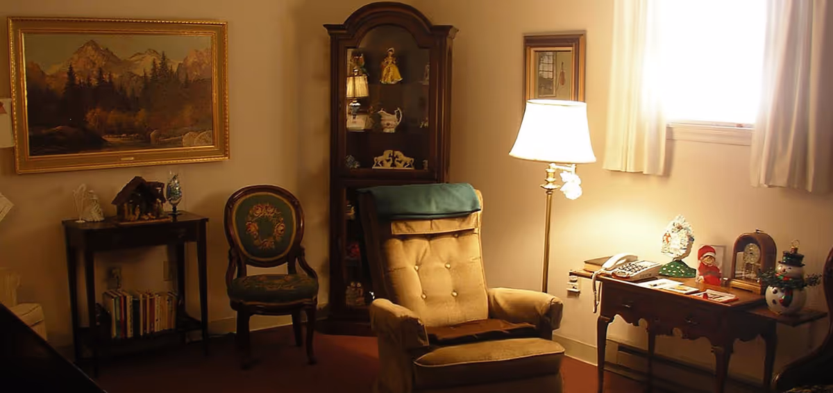 A cozy living room area with a cushioned armchair, a wooden side table with a lamp, a vintage chair with floral upholstery, a small bookshelf with books, a glass display cabinet with decorative items, and a framed landscape painting on the wall.