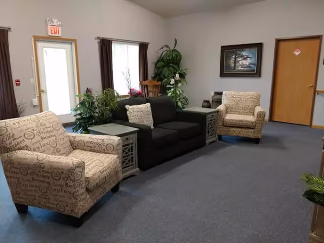 A living room area in a senior living facility with two patterned armchairs and a dark sofa arranged around two side tables. There are several green plants, a window with brown curtains, a wooden rocking chair, a framed landscape painting on the wall, and a door with an exit sign above it.