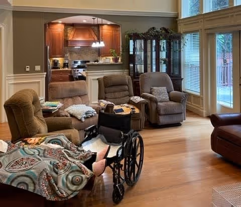 Sunny living room with several recliner chairs, a wheelchair, wooden floors, and an open view into a kitchen.