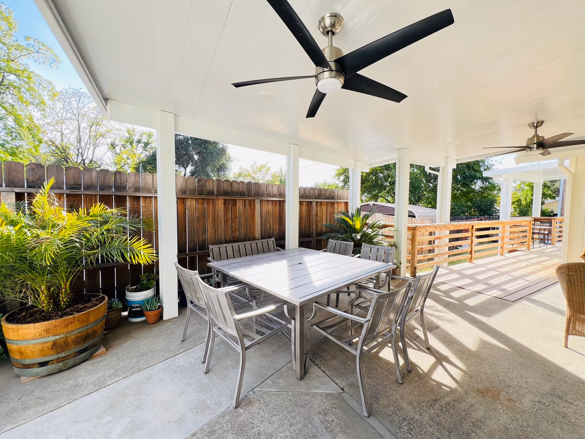 Covered outdoor patio with a metal dining table and chairs, ceiling fans, potted plants, and a wooden fence and railing.