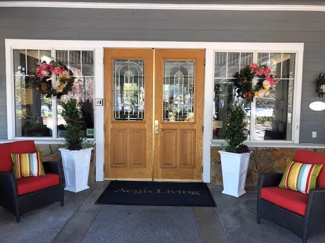 Entrance to Aegis of Carmichael featuring double wooden doors with decorative glass panels, flanked by two large windows each adorned with a floral wreath. Two tall white planters with green plants are positioned on either side of the doors. There are two red cushioned chairs with striped pillows on each side of the entrance. A black doormat with the text 'Aegis Living' is placed in front of the doors.
