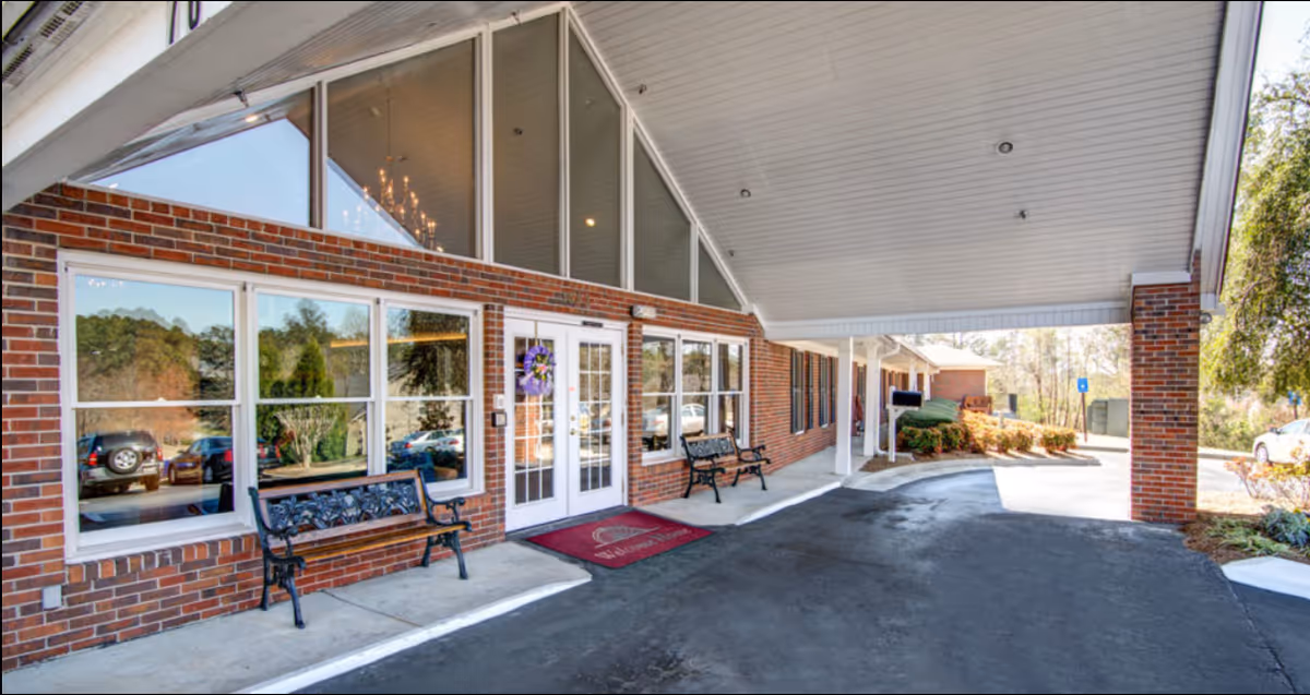 Entrance area of Oaks at Conyers facility showing a covered drop-off zone with brick walls, large windows, two black metal benches, a red welcome mat, and a glass door with a wreath. There are cars parked outside and some greenery around the building.