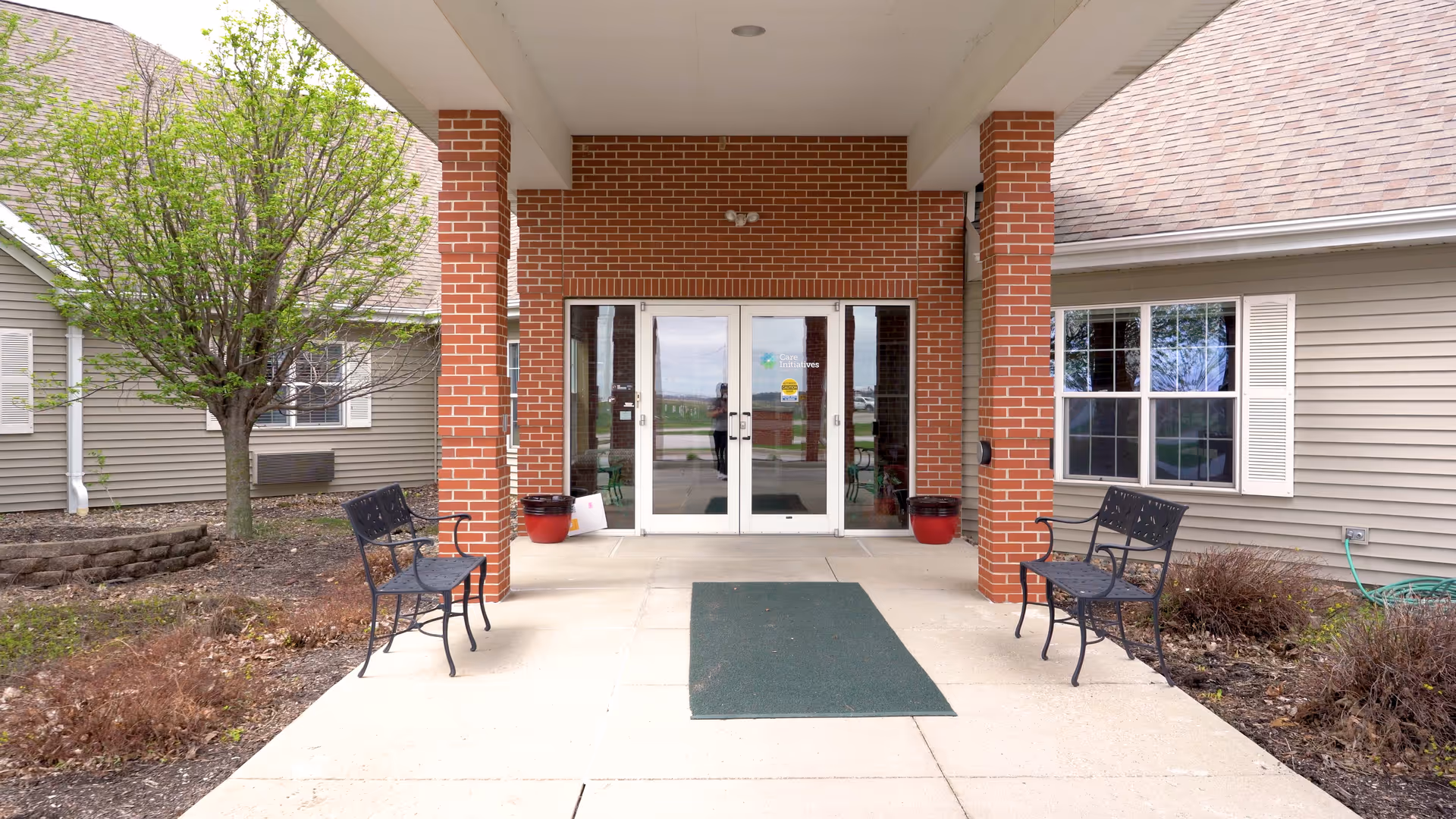 Covered entrance to Avoca Specialty Care with double glass doors framed by brick columns, benches, potted plants, and a green entry mat.
