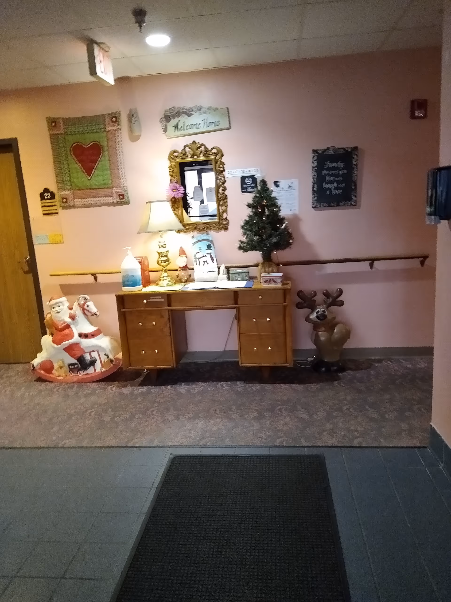An interior hallway area with a wooden desk against a pink wall. On the desk are a lamp, a small decorated Christmas tree, and various decorative items. Above the desk is a gold-framed mirror and a sign that says 'Welcome Home'. To the left of the desk is a rocking horse with a Santa Claus figure, and to the right is a reindeer decoration. The floor has a patterned carpet and a black mat near the foreground.