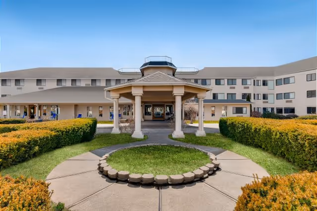 Exterior view of a multi-story senior living facility with a covered entrance, surrounded by neatly trimmed bushes and a circular walkway in the foreground under a clear blue sky.