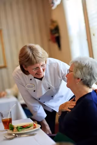 A staff member wearing a white uniform and name tag is smiling and leaning towards an elderly woman seated at a dining table. The table has a plate with a sandwich and a glass of iced tea. The setting appears to be a dining area with soft lighting and window blinds in the background.