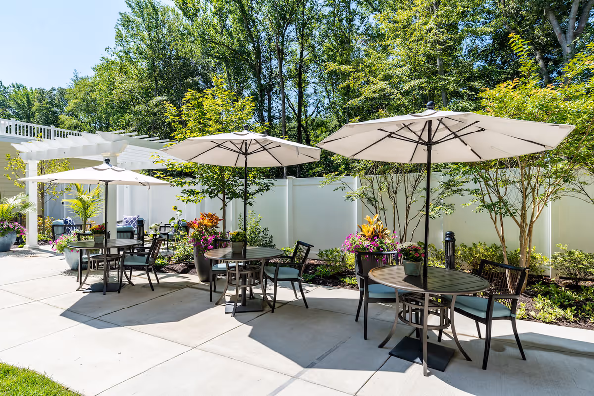 Outdoor patio area with round tables and chairs under large white umbrellas, surrounded by potted plants and greenery, with a white fence and trees in the background under a clear blue sky.