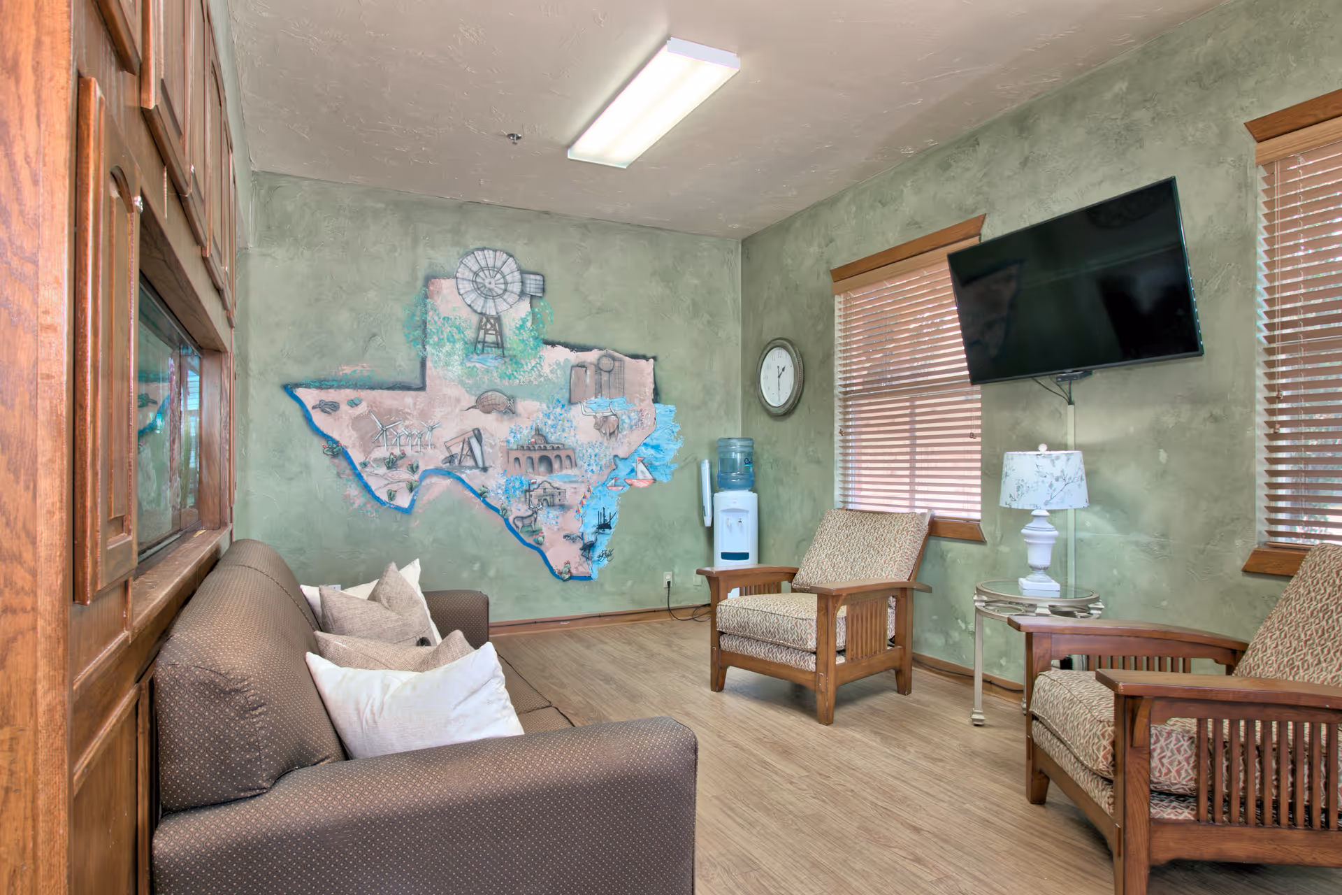 A cozy living room with a brown couch and two wooden armchairs with patterned cushions. The walls are painted green with a mural of the state of Texas featuring various landmarks. There is a water cooler, a wall clock, a flat-screen TV mounted on the wall, and a small round table with a white lamp. The room has wooden blinds on the windows and wood flooring.