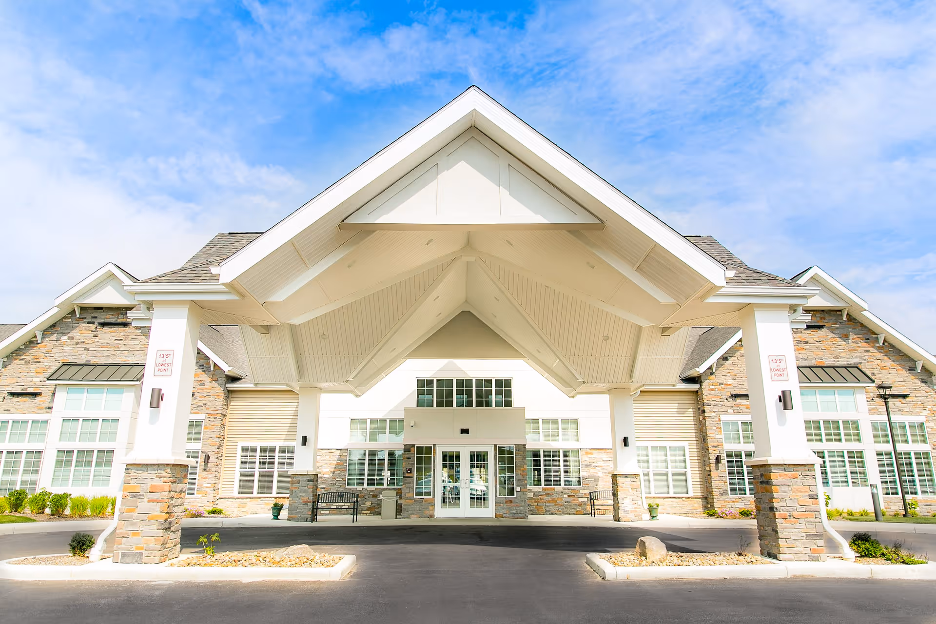 Front exterior view of a senior living facility with a large covered entrance supported by stone and white pillars, multiple windows, and a clear blue sky above.