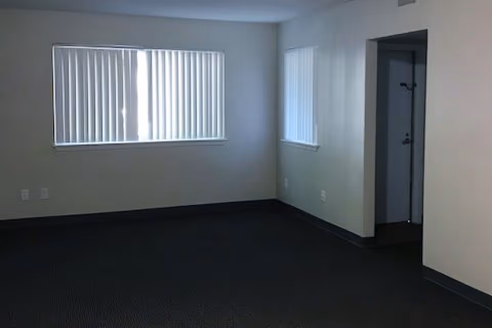 Empty room with dark carpet, beige walls, two windows with vertical blinds, and an open doorway leading to another area.