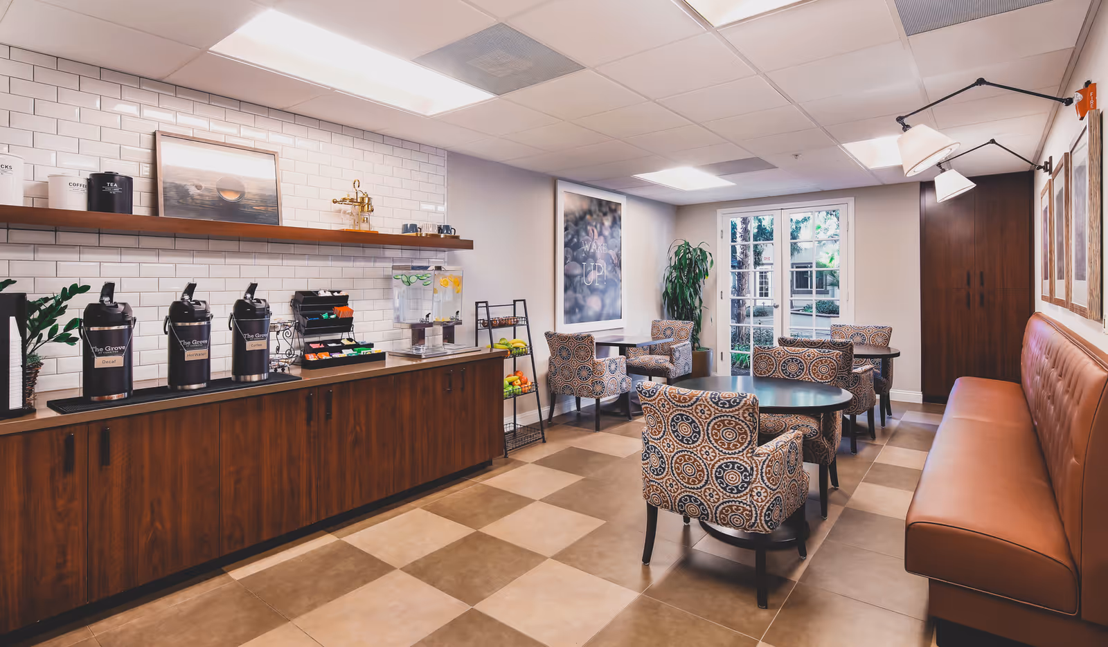 Bright communal lounge with a coffee station along a tiled wall, patterned armchairs, round tables and a leather banquette.