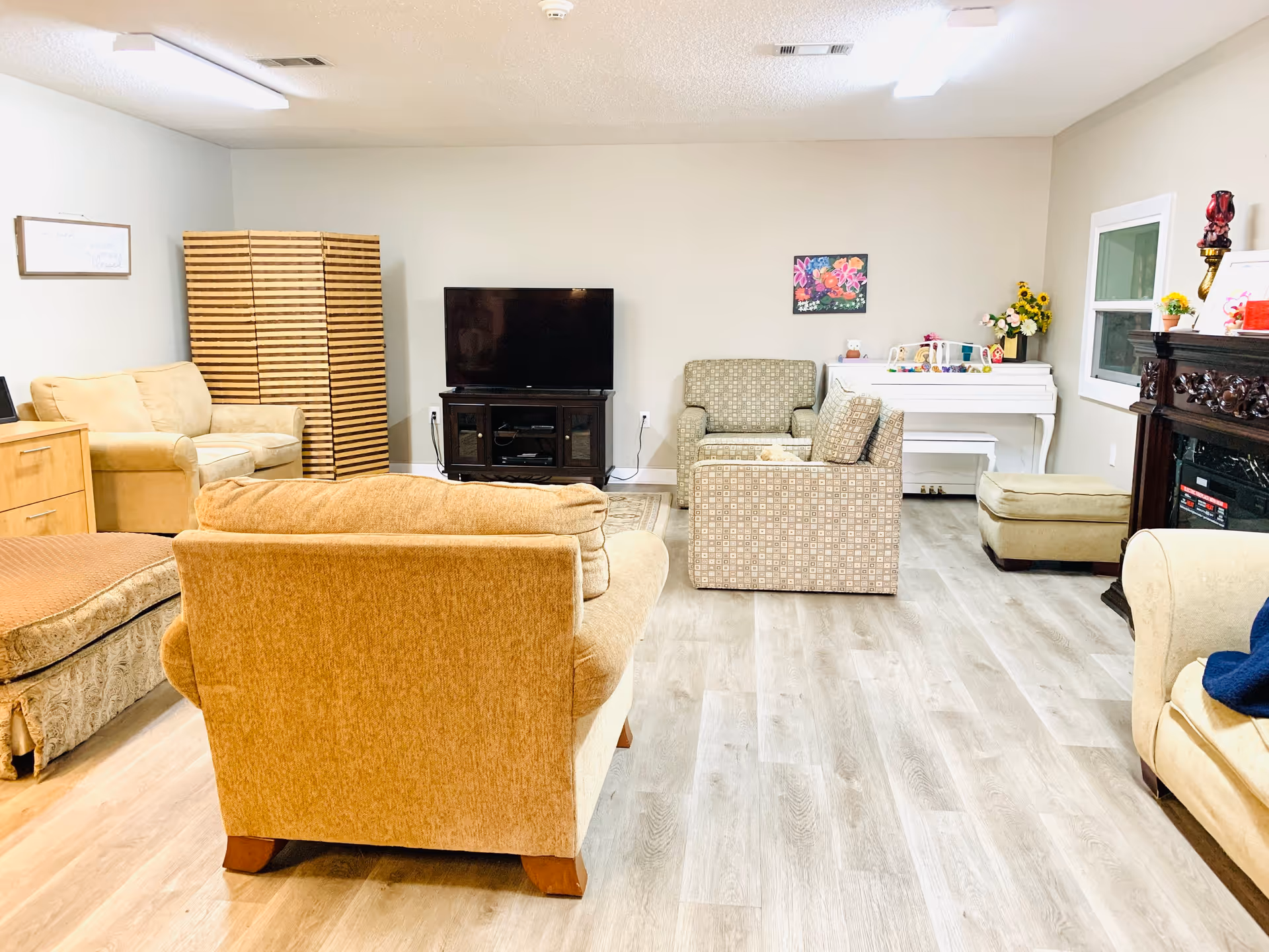 A cozy living room with beige and patterned armchairs, a large flat-screen TV on a dark wooden stand, a white piano with decorative items on top, a wooden room divider, and a fireplace with a dark wooden mantel. The room has light-colored wood flooring and neutral-colored walls.