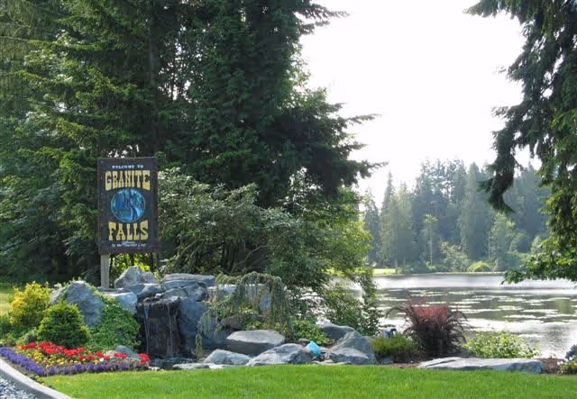 A landscaped outdoor area featuring a welcome sign for Granite Falls surrounded by rocks, flowers, and greenery with a lake and trees in the background.