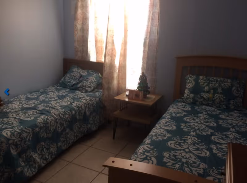 A small bedroom with two single beds placed parallel to each other. Both beds have matching teal and white patterned bedspreads and pillows. Between the beds is a small wooden nightstand with a miniature Christmas tree and a framed photo on it. A window with light-colored patterned curtains is behind the nightstand, allowing natural light to enter the room. The floor is tiled.