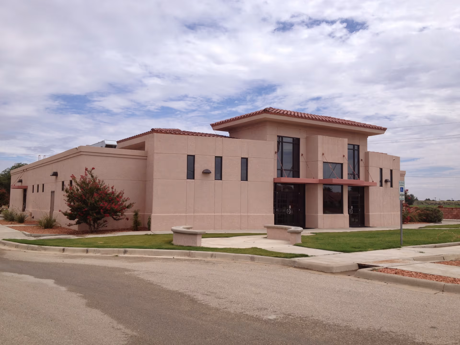 Two-story stucco building with a tiled roof and glass double-door entrance set in landscaped grounds under a cloudy sky.