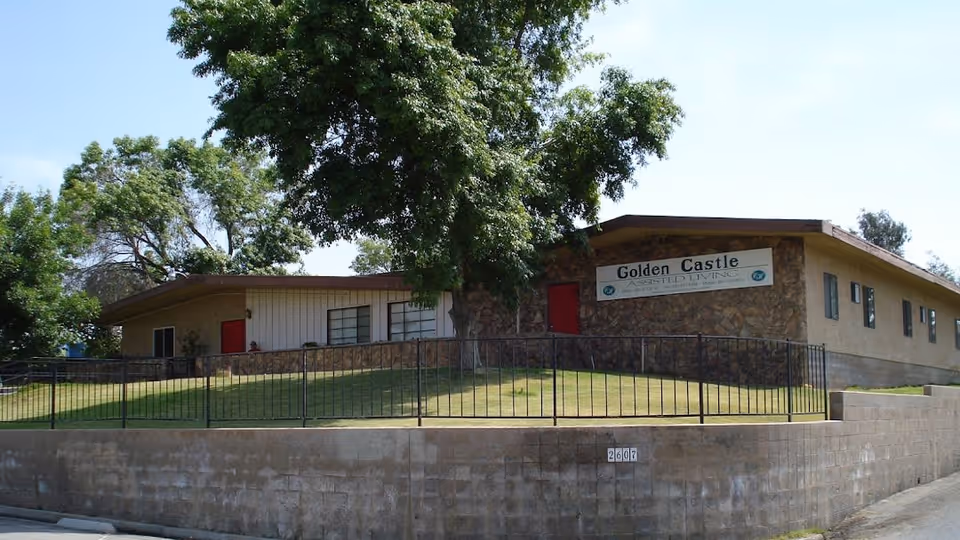 Exterior view of Golden Castle Assisted Living facility, a single-story building with stone and beige walls, red doors, several windows, a black metal fence, a grassy area, and large trees under a clear blue sky.