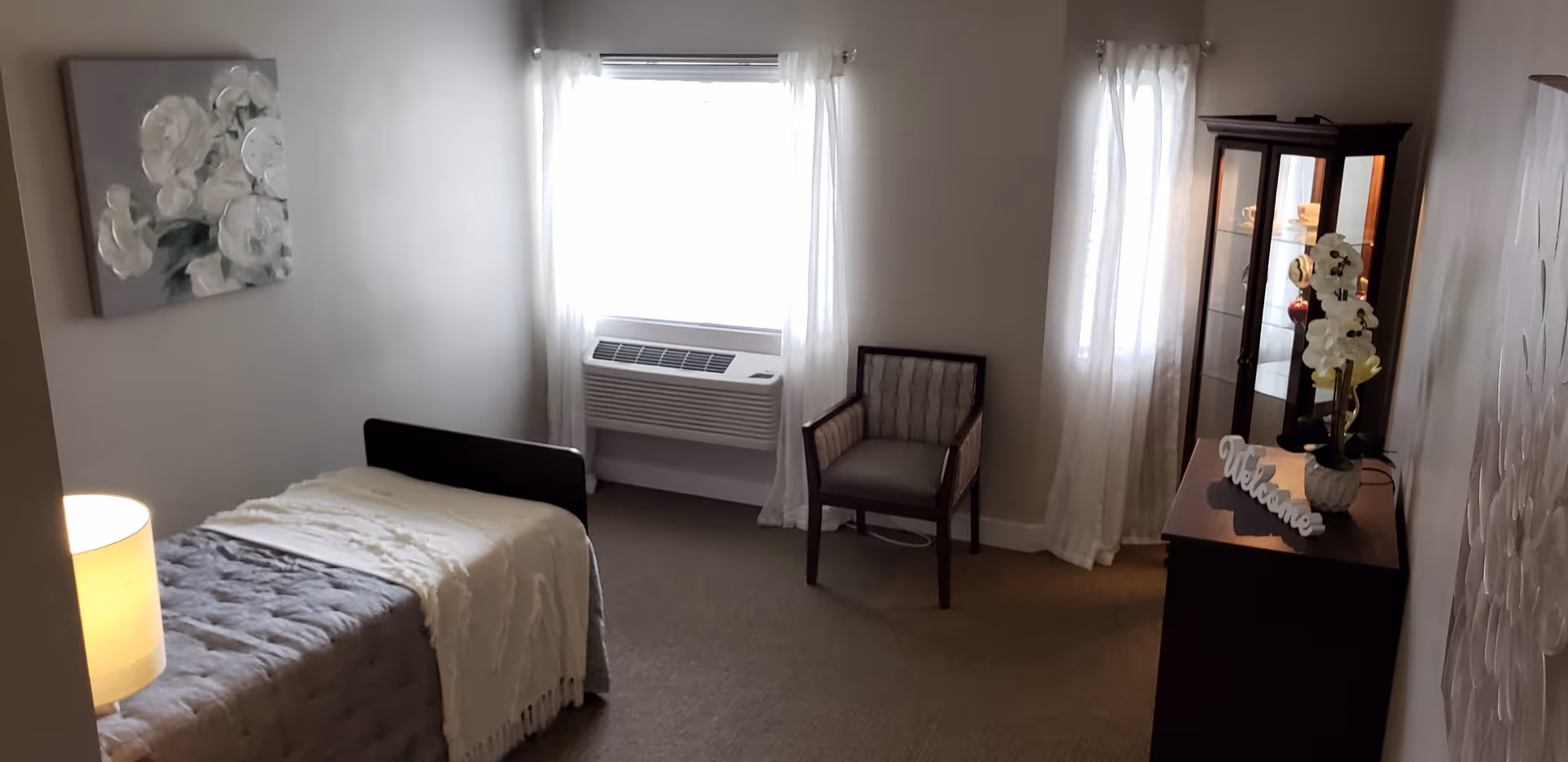 A small, cozy bedroom with a single bed covered in a gray quilt and a white throw blanket. There is a lamp on a bedside table next to the bed. The room has two windows with sheer white curtains, a wall-mounted air conditioning unit below one window, a wooden chair with cushioned seat and backrest, a wooden cabinet with glass doors displaying decorative items, and a 'Welcome' sign on top. The walls are light-colored and decorated with floral artwork.