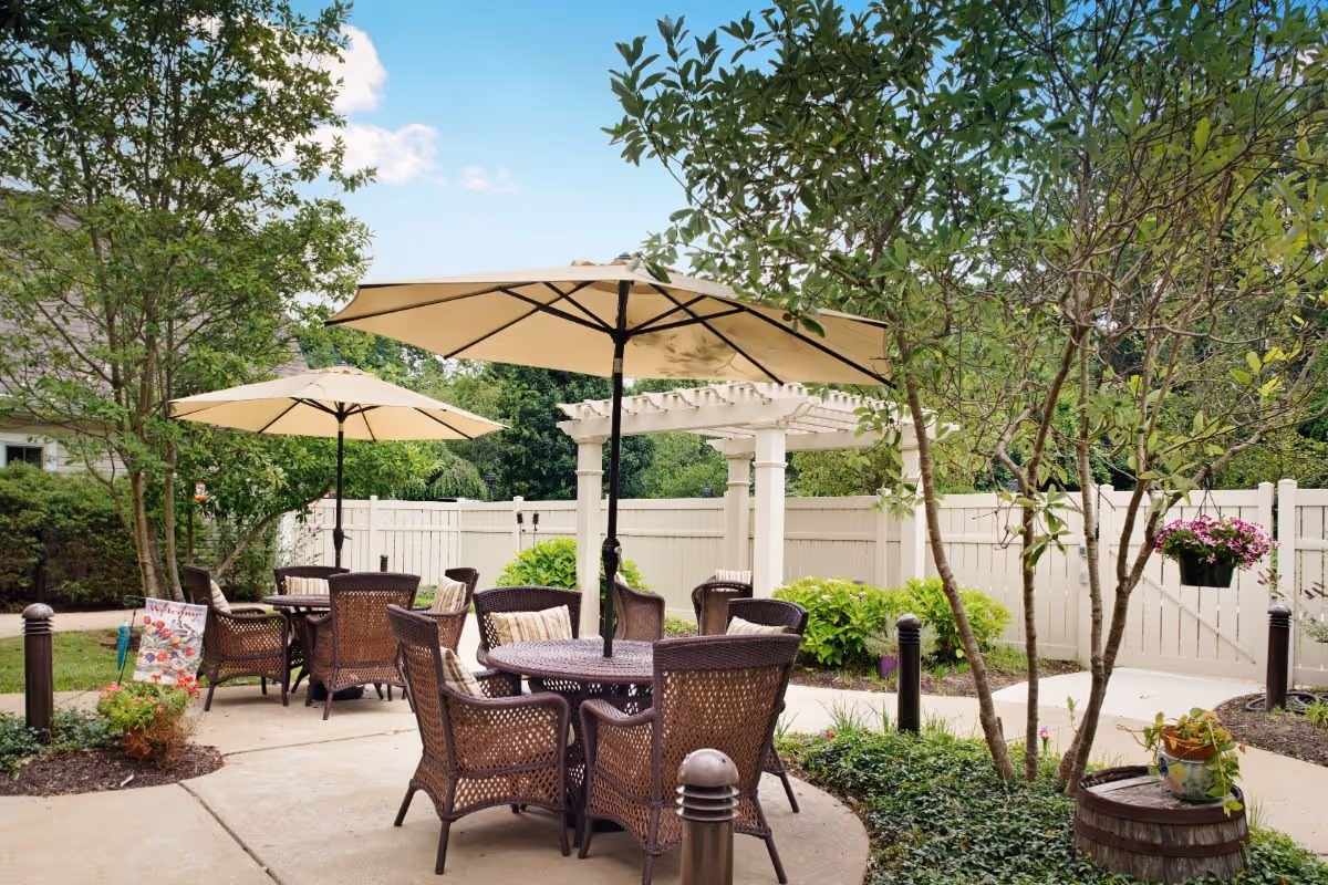 Outdoor patio area with round wicker tables and chairs, each table shaded by a large beige umbrella. The patio is surrounded by greenery, including trees, bushes, and potted plants. A white fence and a white pergola are visible in the background under a blue sky with some clouds.