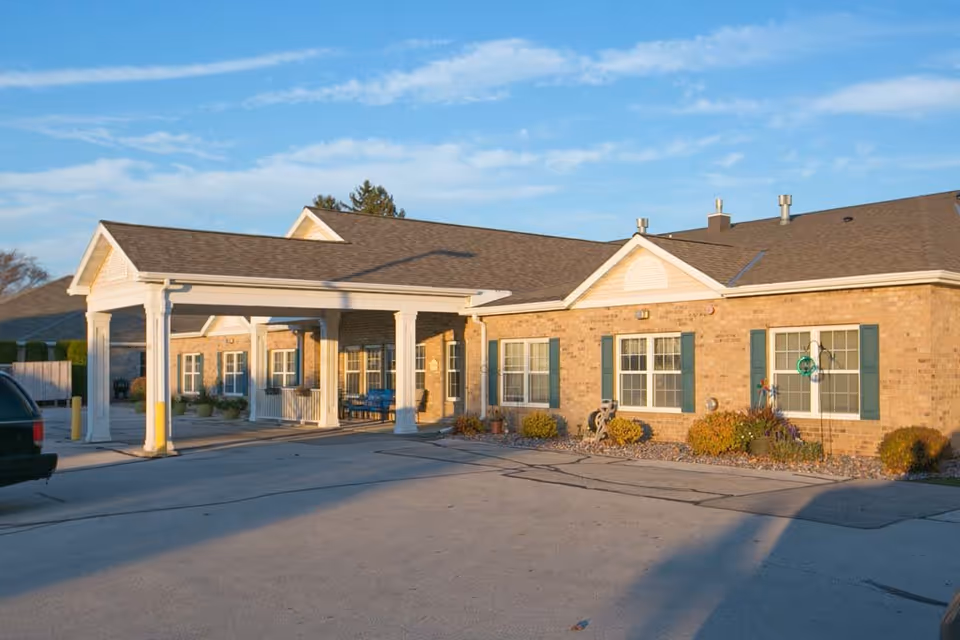 Exterior view of Harbor View Assisted Living facility showing a single-story brick building with green window shutters, a covered entrance supported by white columns, and a paved driveway in front under a blue sky with some clouds.