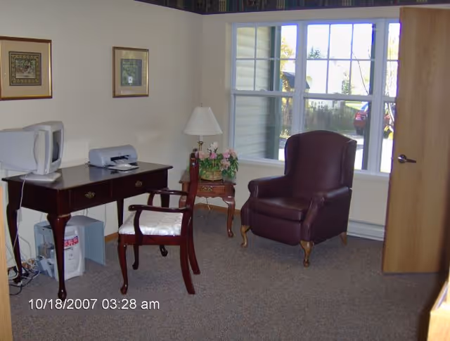 A small room with a wooden desk holding a computer monitor and a printer, a wooden chair with a cushioned seat, a side table with a lamp and a flower pot, and a brown leather armchair near a window showing an outdoor view.
