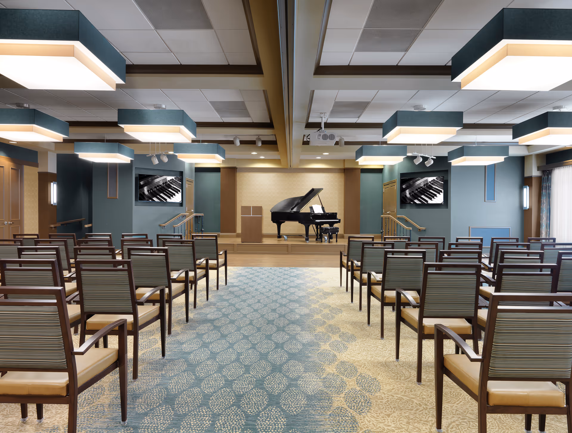 A large event room with rows of chairs facing a stage with a grand piano and wall-mounted screens.