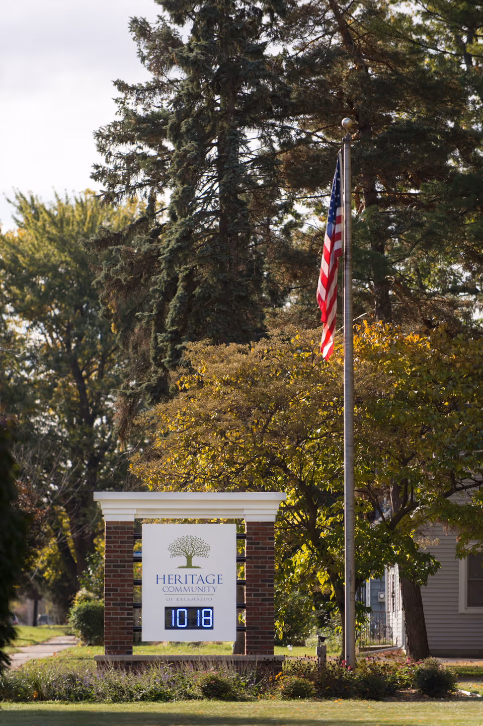 Outdoor view of a sign for Heritage Community of Kalamazoo with a digital clock displaying 10:18. The sign is supported by brick pillars and is surrounded by greenery, trees, and a flagpole with an American flag.