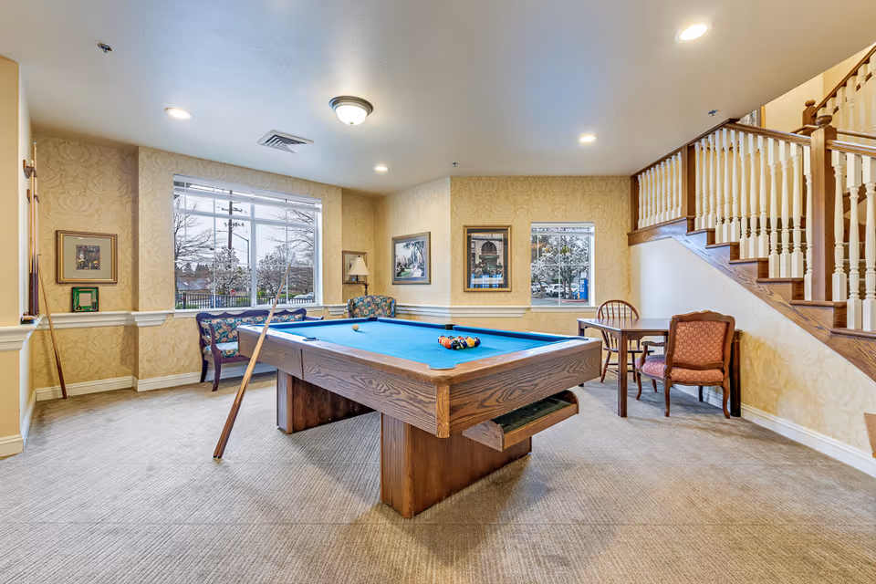 Bright common room with a pool table, seating, windows, and a staircase.