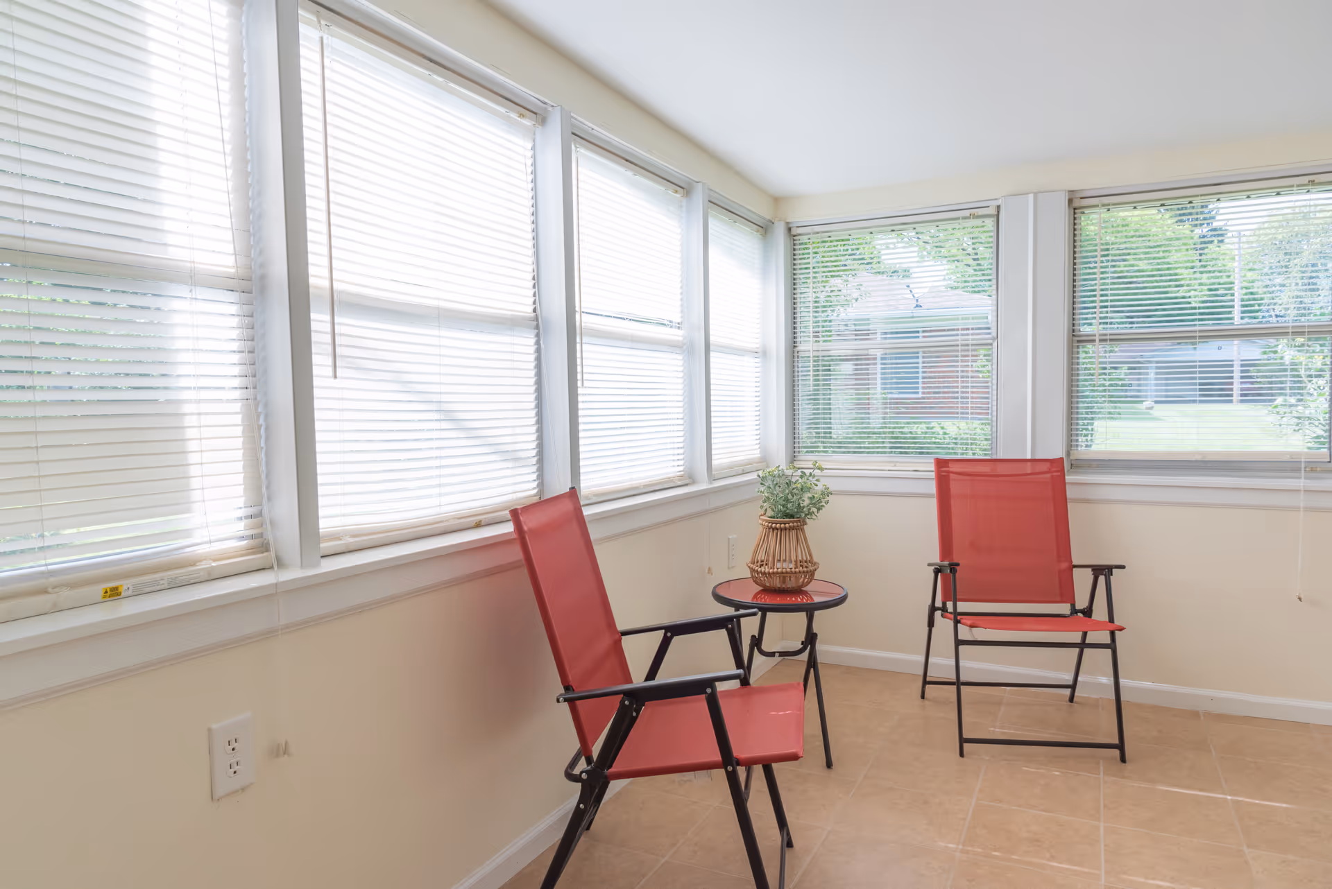 A bright sunroom with large windows covered by white blinds. The room contains two red mesh chairs and a small round table with a decorative plant in a wicker vase. Outside the windows, greenery and parts of neighboring buildings are visible.