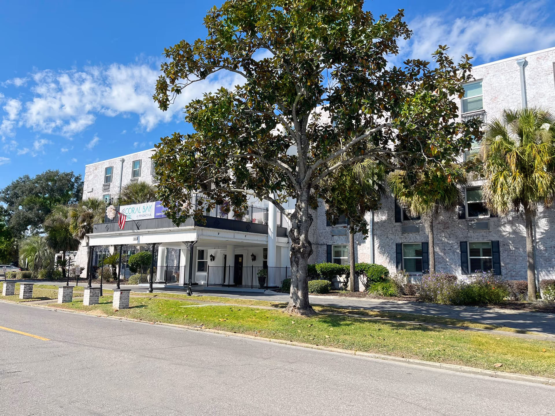 Exterior view of Coral Bay at Pensacola senior living facility showing a three-story white brick building with multiple windows, a covered entrance with columns, a large tree in front, and a clear blue sky with some clouds.