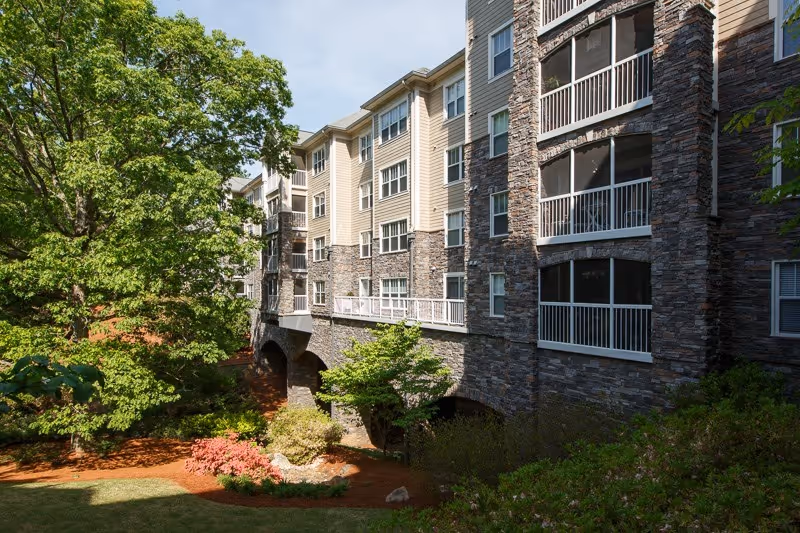 Exterior view of a multi-story senior living building with stone facade, screened balconies, and landscaped trees and shrubs.