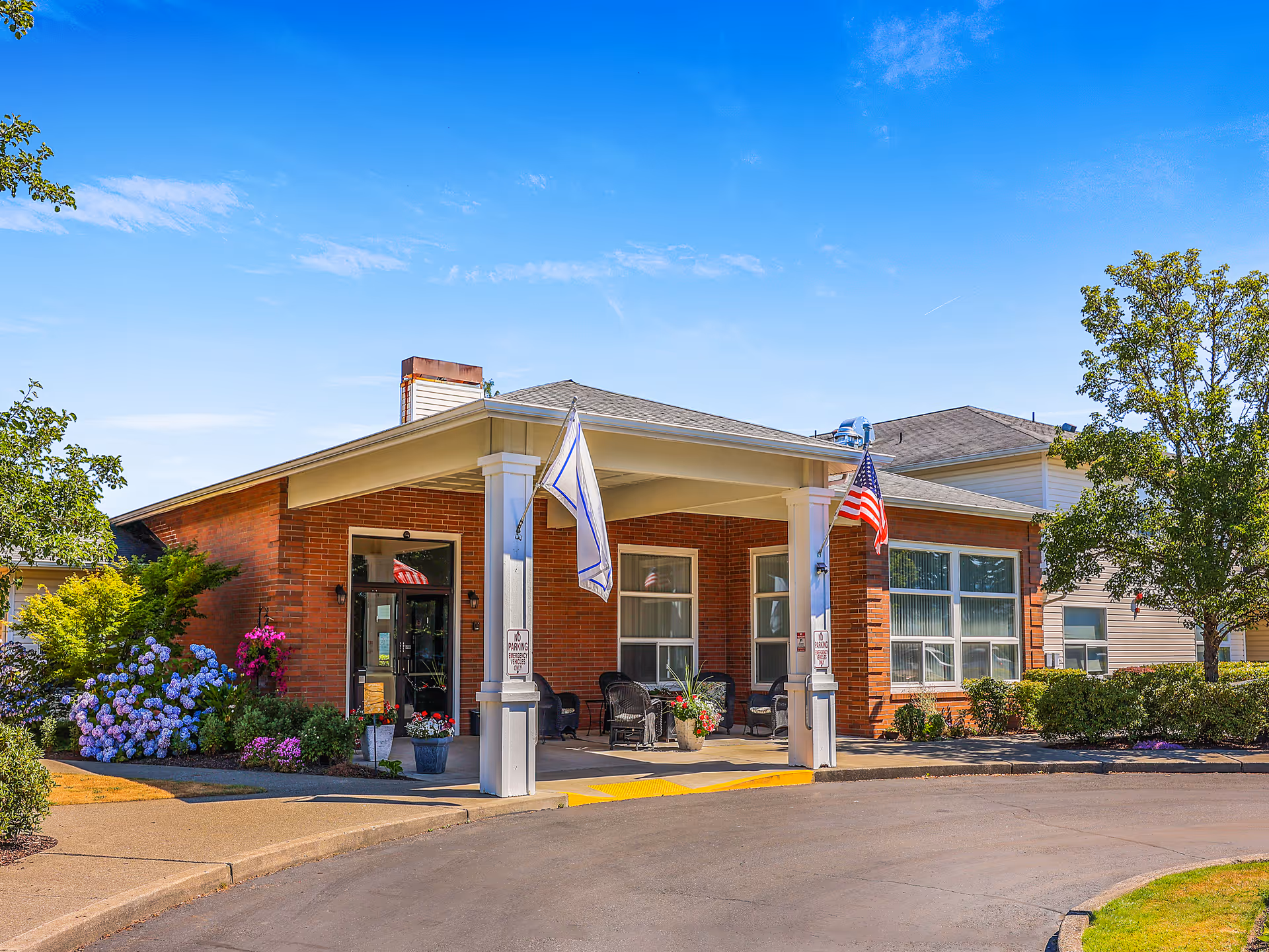 Entrance of a brick assisted living facility with a covered porte-cochere, flags, potted plants, and outdoor seating.