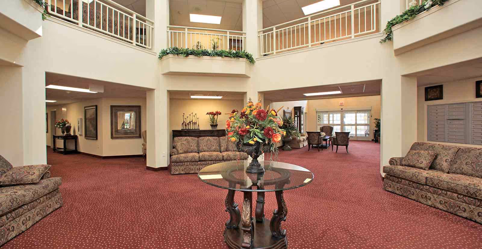 Spacious carpeted senior living lobby with sofas, a central glass-top table holding a floral arrangement, and a second-floor railing balcony.