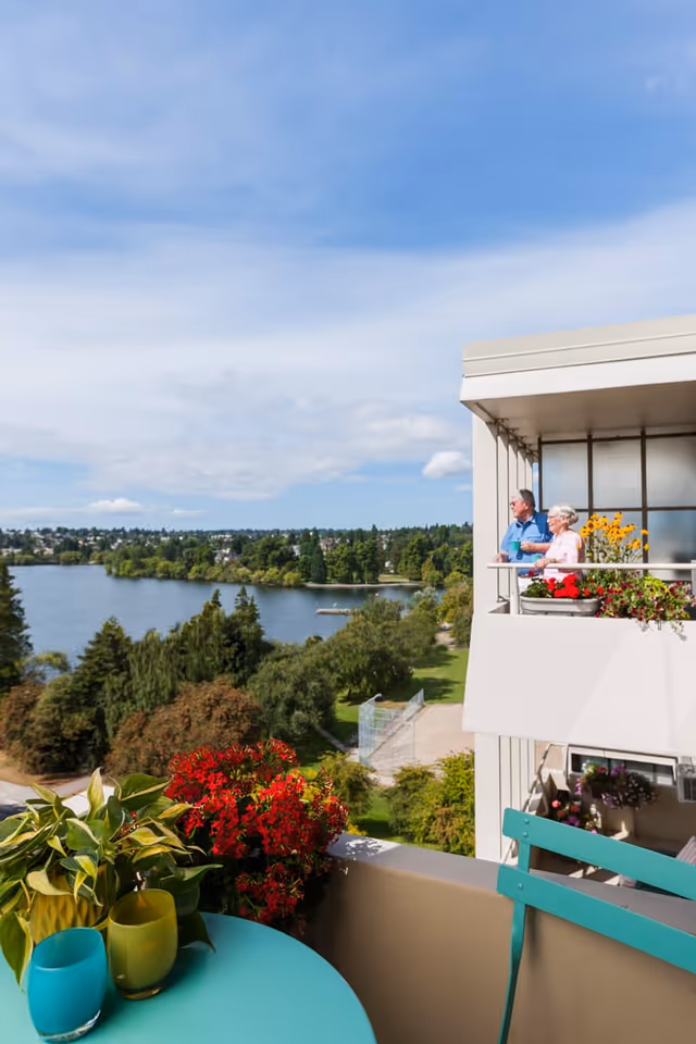 View from a balcony overlooking a lake and green trees under a blue sky. Two elderly people stand on a neighboring balcony with flower pots, enjoying the scenery. In the foreground, there is a turquoise table with colorful glasses and a plant, along with a turquoise chair.