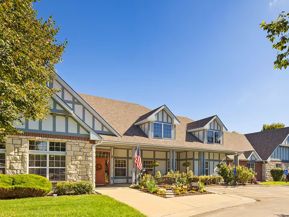 Exterior view of The Windsor of Lawrence facility showing a large building with stone and wood paneling, multiple windows, a front porch with an American flag, landscaped garden beds, and a clear blue sky.