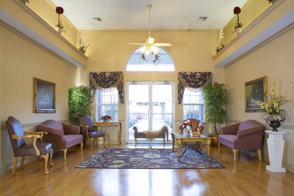 A bright and elegant nursing center lobby with polished wooden floors, a floral area rug, and multiple seating options including armchairs and sofas. The room features large windows with floral valances, potted plants, framed paintings on the walls, a ceiling fan with light, and a glass double door entrance letting in natural light.