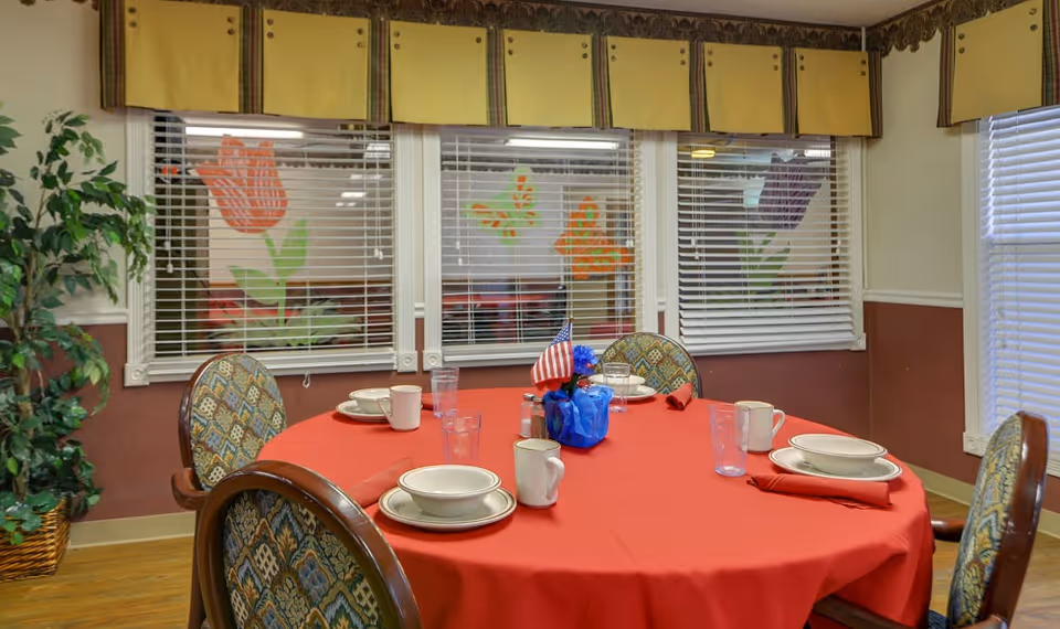 Round dining table covered with a red tablecloth set with plates, cups and an American flag centerpiece in a decorated dining room with windows and blinds.