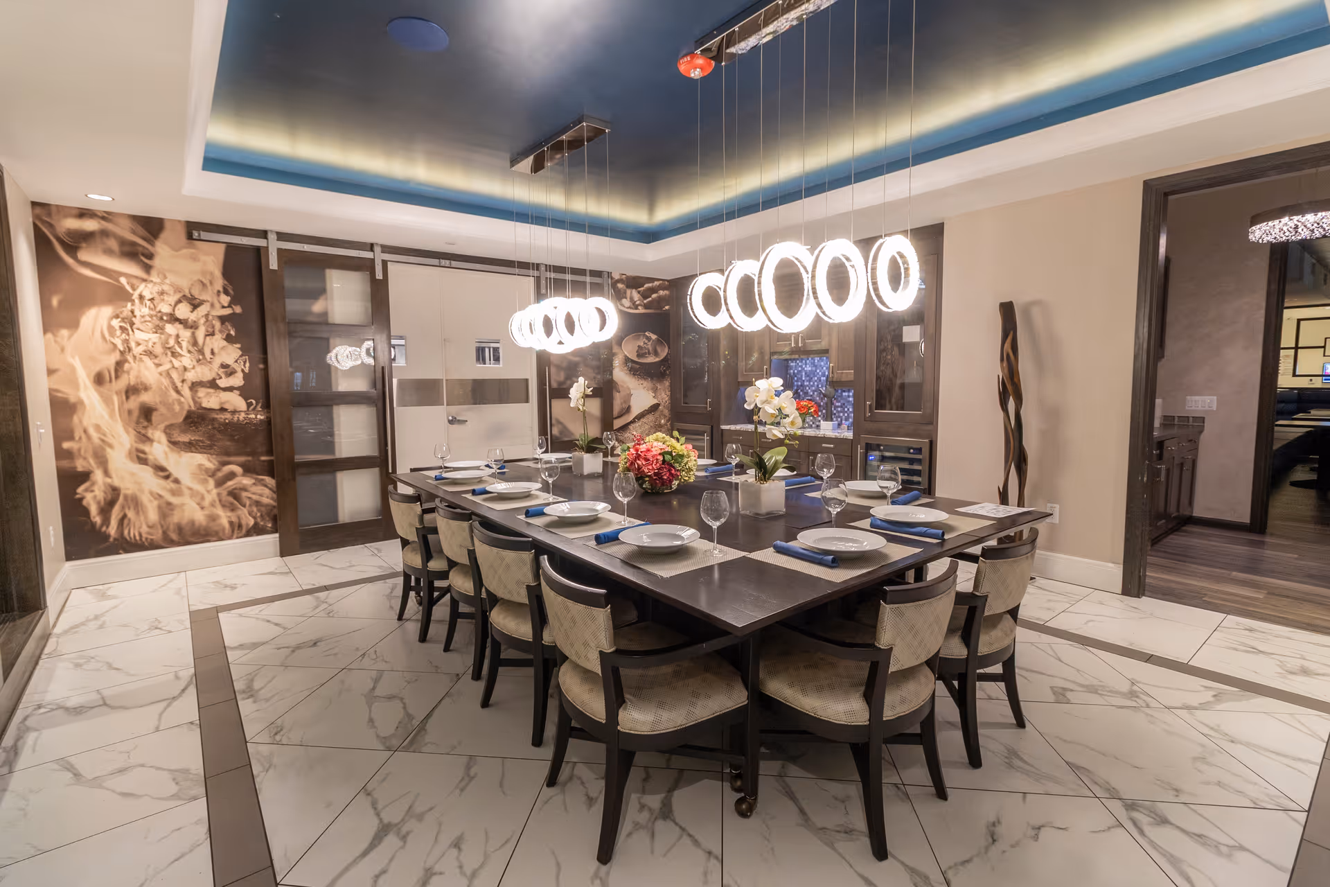 A modern dining room with a large square table set for twelve people. The table has white plates, wine glasses, and blue napkins. Above the table hang two contemporary circular light fixtures. The room features marble tile flooring, a blue ceiling with recessed lighting, and decorative wall art depicting food. There are glass-paneled sliding doors and a doorway leading to another room.