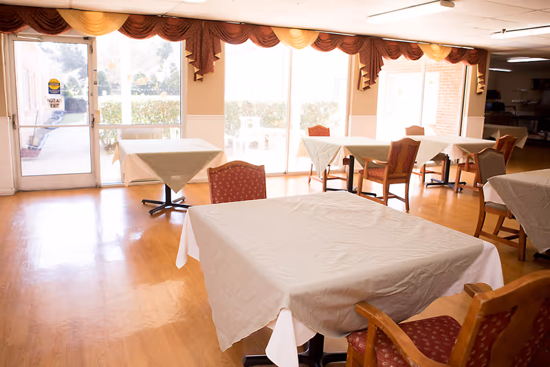 A dining room with several tables covered with white tablecloths and surrounded by wooden chairs with red patterned cushions. Large windows with decorative valances allow natural light to fill the room, and a door with a 'Not an Exit' sign is visible on the left side.