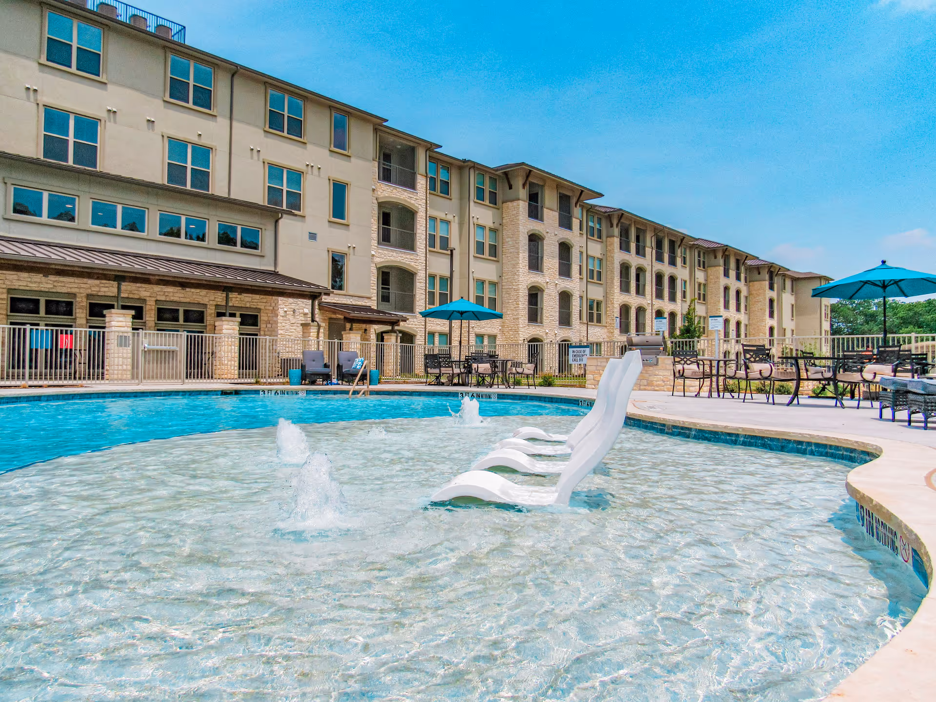 Outdoor swimming pool area at Preserve at Willow Park with clear blue water, white lounge chairs partially submerged in the shallow end, water fountains, patio tables with umbrellas, and a multi-story residential building in the background under a clear blue sky.