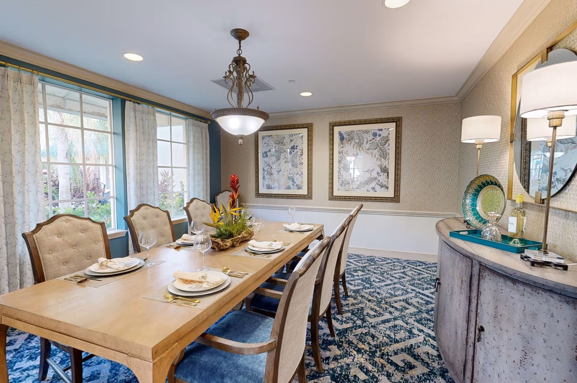 A well-lit dining room with a long wooden table set for six people. The table has plates, napkins, wine glasses, and a centerpiece with tropical flowers. The room features large windows with curtains, two framed floral artworks on the wall, a patterned blue carpet, and a sideboard with decorative items and a lamp.