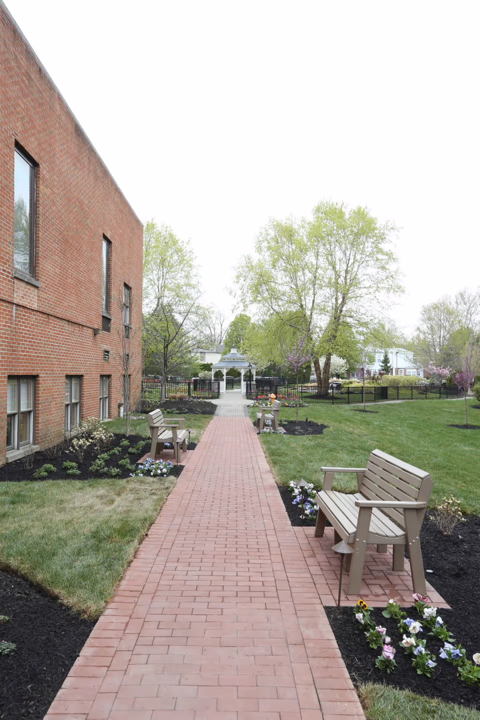 A brick pathway lined with benches and flower beds leads through a garden area beside a red brick building. Trees with fresh green leaves and a white gazebo are visible in the background under an overcast sky.