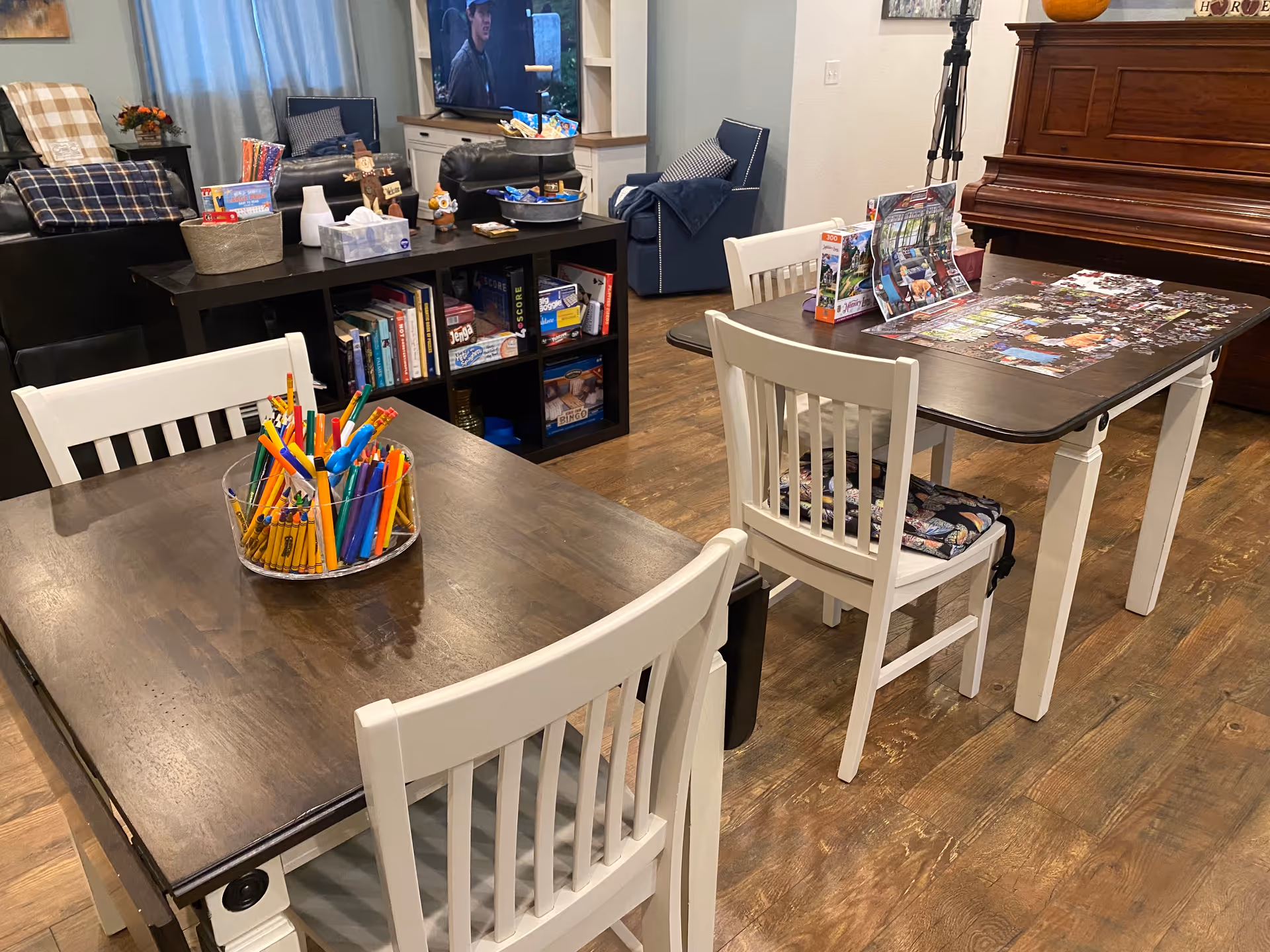 A cozy senior living common area with two dark wooden tables and white chairs. One table has a clear container filled with colorful pencils and markers, while the other table has a partially completed puzzle and puzzle boxes. In the background, there is a bookshelf with board games and books, a television showing a person, and comfortable seating including a blue armchair and a black leather couch. A piano is visible on the right side of the room.