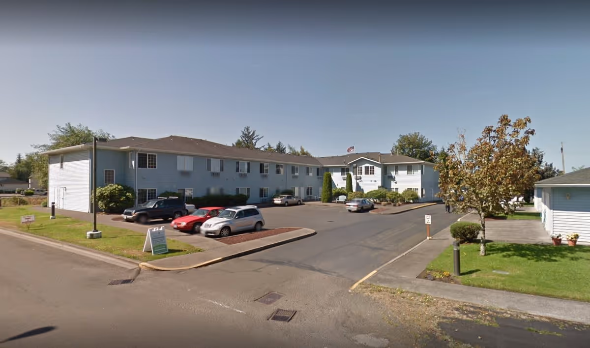 Exterior view of a two-story light blue senior living facility building with a parking lot in front containing several parked cars. There is a tree and some bushes on the right side, and the sky is clear.